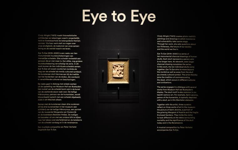On a black wall, there is an exhibition caption with the title ‘Eye to Eye’ and a small piece of art hanging there.