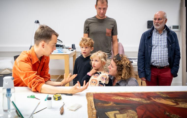 Iemand geeft uitleg aan een gezin bij een schilderij dat op tafel lifgt.