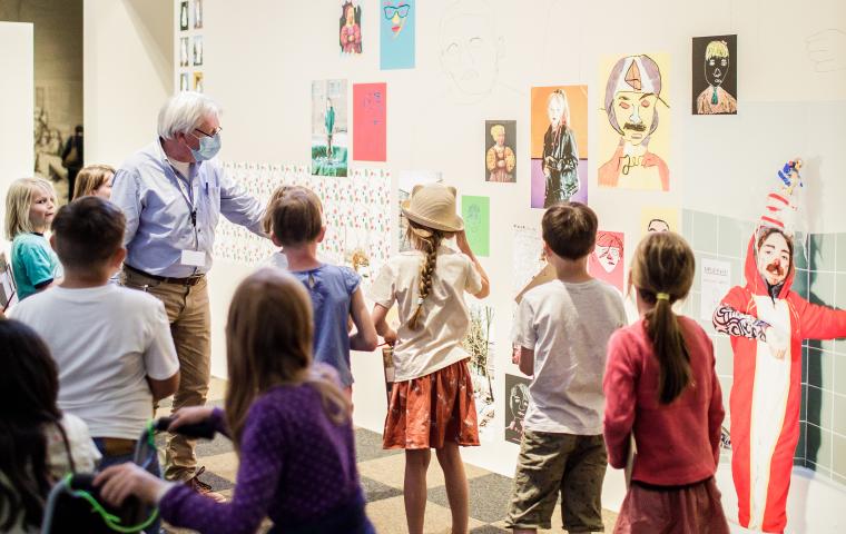 Een groep kinderen kijkt naar een muur gevuld met kindertekeningen.