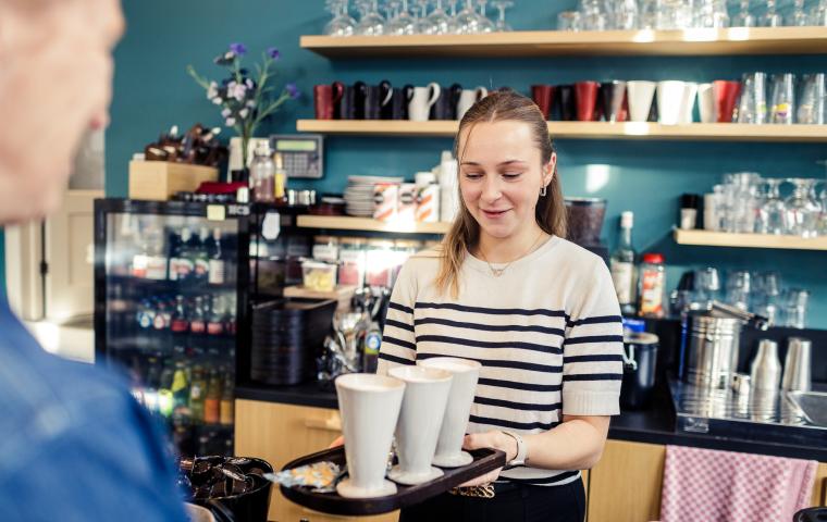 Iemand glimlacht en overhandigt een plateau met 3 kopjes koffie.