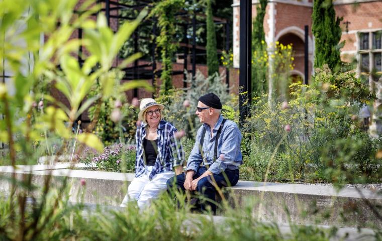Twee mensen zitten al pratend en lachend in een tuin, enkele weelderige planten op de voorgrond.