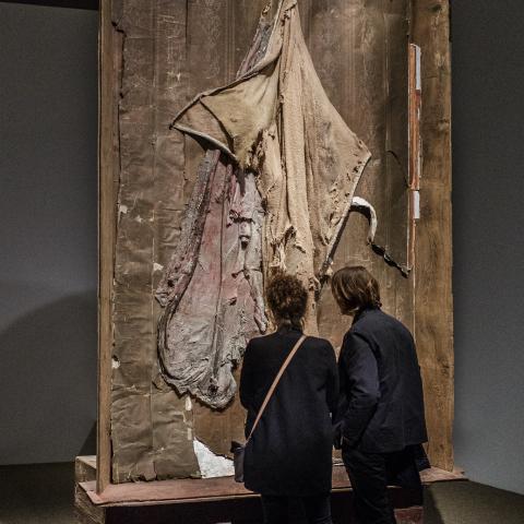 Twee bezoekers kijken naar een groot hedendaags kunstwerk, gemaakt uit textiel en een houten kader.