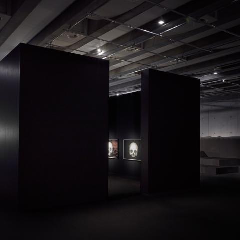 In a dark room, the walls are arranged in a cube; two illuminated drawings are visible through an opening.