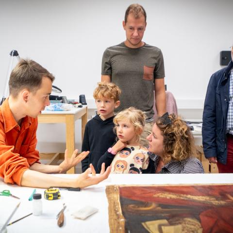 Iemand geeft uitleg aan een gezin bij een schilderij dat op tafel lifgt.