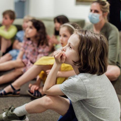 Een groep kinderen zit op een tapijt en kijkt allemaal dezelfde richting uit.