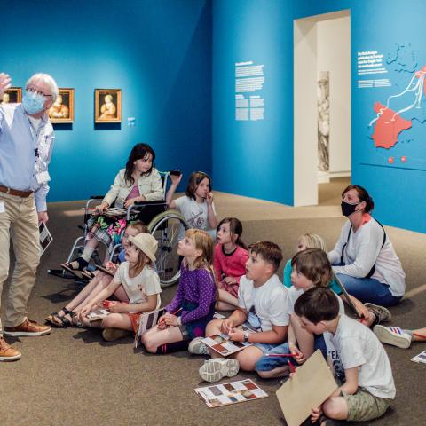 Een groep kinderen zit op het tapijt van een museumzaal en kijkt naar een gids met mondmasker.