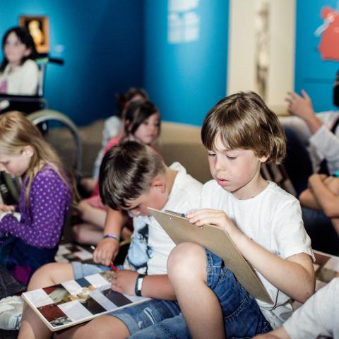 Een groep kinderen zit in een museumzaal en leest brochures.