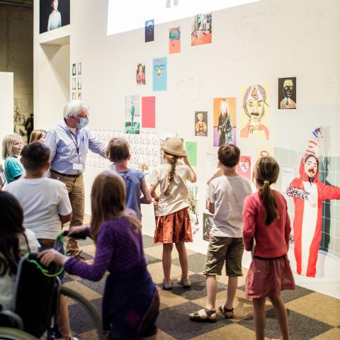 Een groep kinderen kijkt naar een muur gevuld met kindertekeningen.