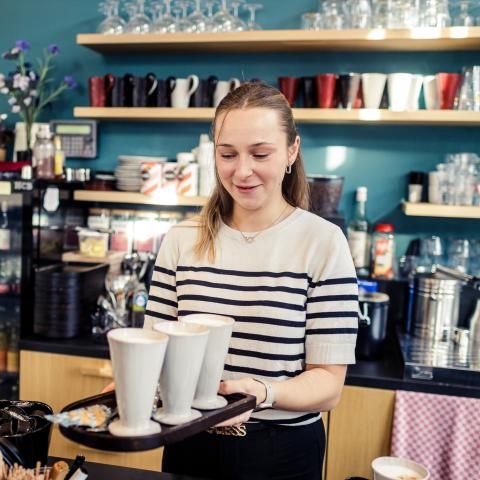 Iemand glimlacht en overhandigt een plateau met 3 kopjes koffie.
