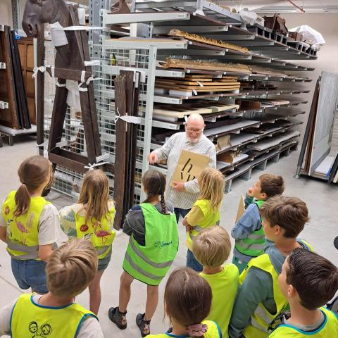 Een groep kinderen luistert naar een gids in een depot.