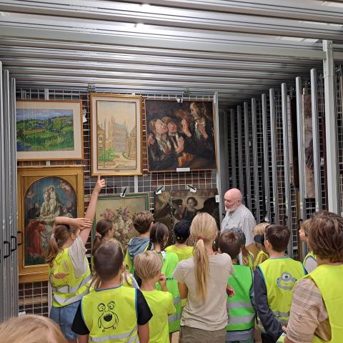 Een groep kinderen staat in een depot gevuld met schilderijen.