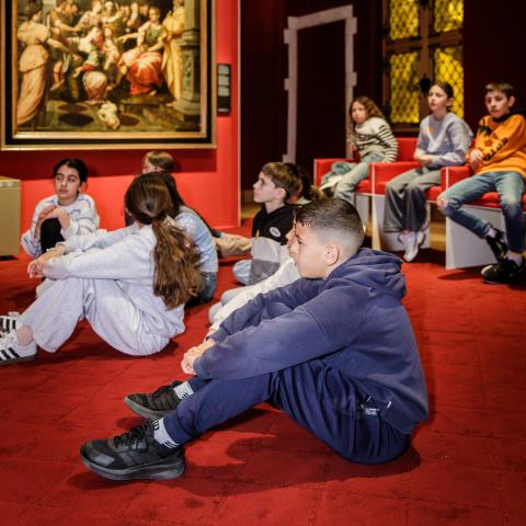 Een groep kinderen zit op een rood tapijt in een museumzaal.