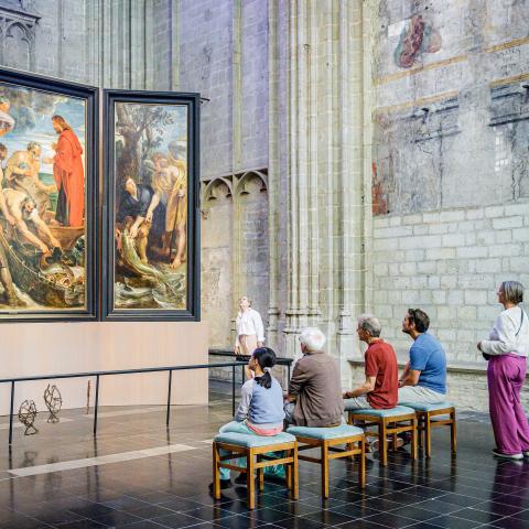 Eine Gruppe von Menschen sitzt auf Bänken vor einem großen Triptychon.