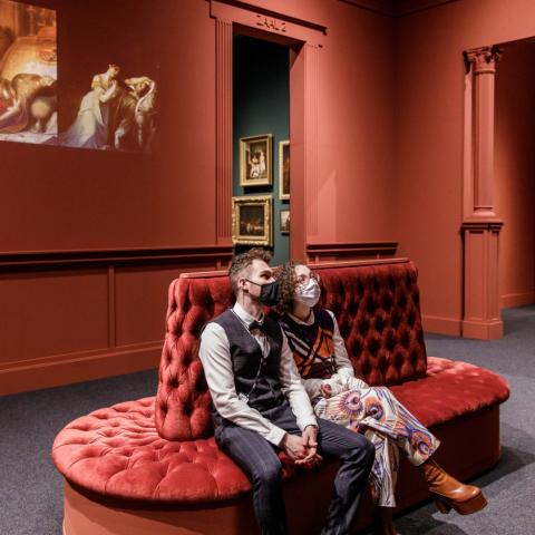 Twee bezoekers met mondmasker zitten op een rode floeren bank in een museumzaal.