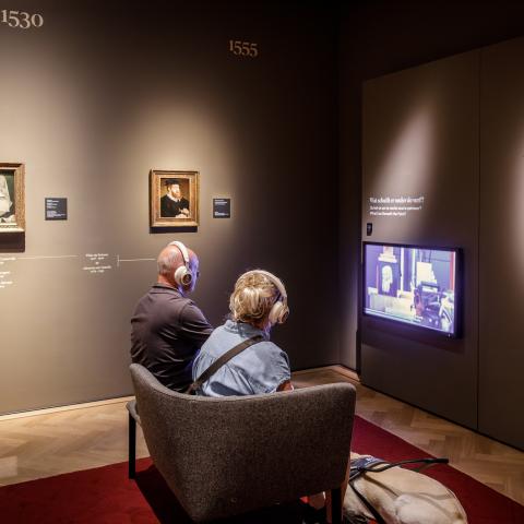 Twee bezoekers zitten in de zetel in een museumzaal met verschillende portretten.