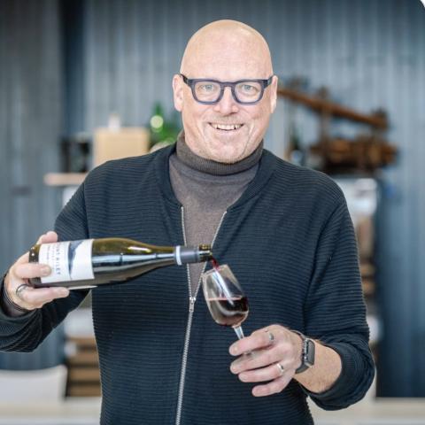 Een man kijkt glimlachend in de camera en schenkt een glas wijn in.